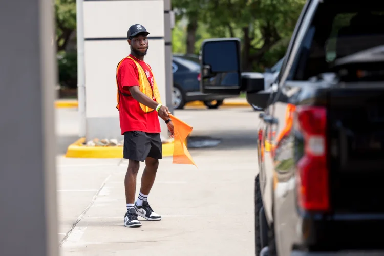 A staff member with neon orange vest and flag in hand directs a pickup truck through a dropoff area in front of a building.