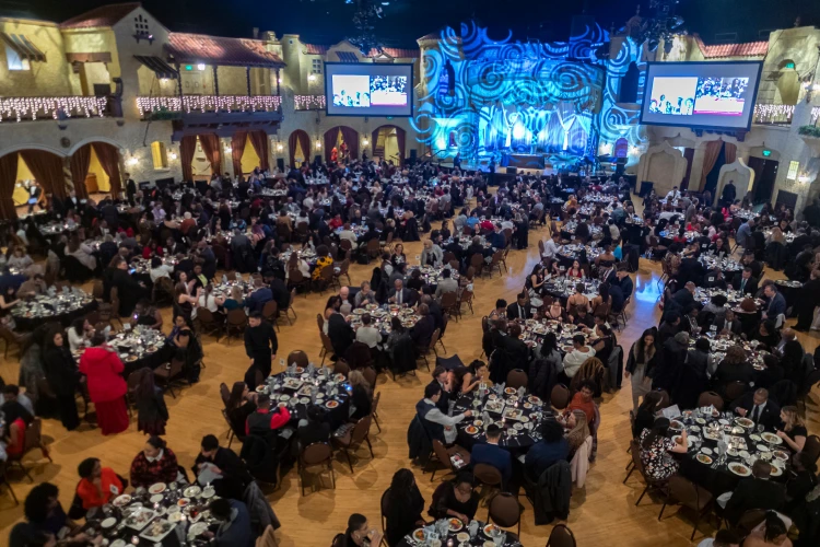 A ballroom is set up with dozens of tables circled by dinner participants. Screens flank a lit stage with blue and white swirls and a podium.