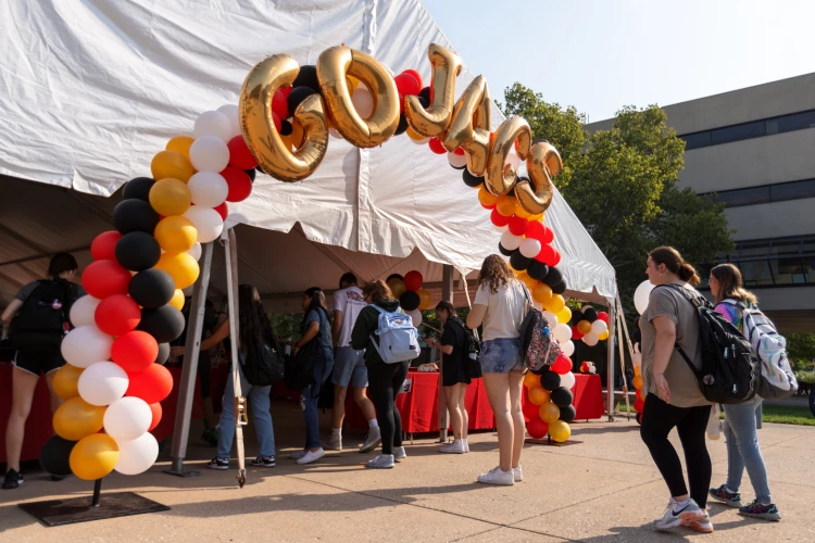 Students line up to enter an outdoor event tent with an arch of red, white, black, and gold balloons with some that spell out Go Jags.