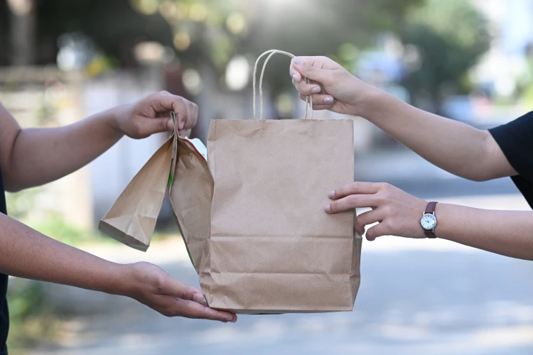 One pair of arms outreached hands three brown paper bags of delivery food to another pair of outstretched arms.