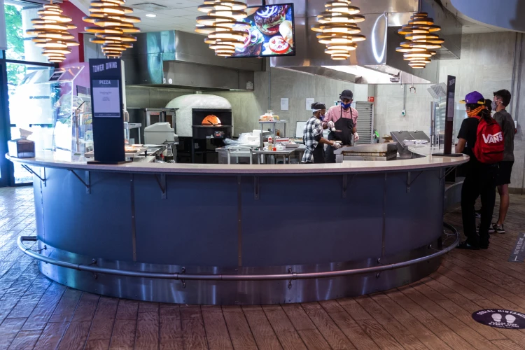 Two students stand at a curved counter to order food; a pizza oven glows in the background among kitchen appliances and equipment.