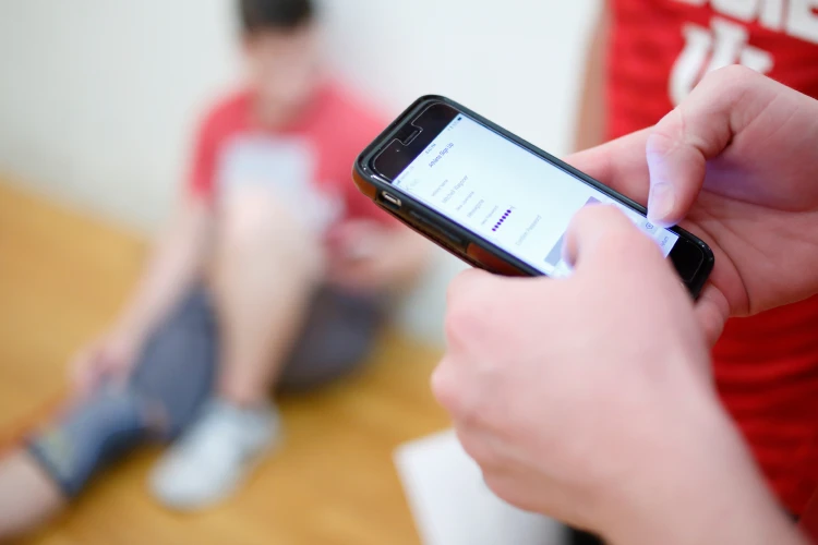 Hands holding a phone type on the screen; two other students in athletic gear are nearby out of focus.