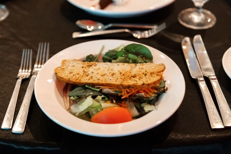 A salad and piece of bread sit in a bowl surrounded by silverware atop a black tablecloth.