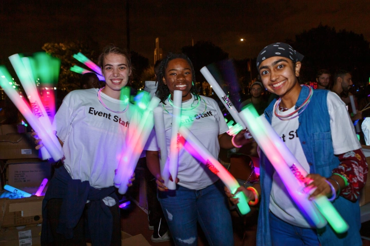 Three people with tee shirts reading Event Staff hold up large glow sticks at an evening event on campus.