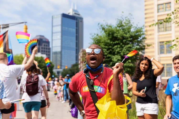 Students walk down a street during Indianapolis’ Pride Parade waving rainbow flags while a spectator speaks as they pass.