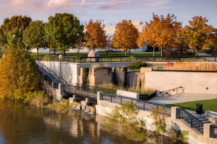 Fall trees are seen along the White River State Park promenade with stairs, a waterfall, and riverfront views.
