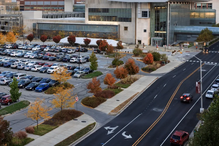A car drives on a road toward the Campus Center, with rows of parked cars in a parking lot to the side of the road.