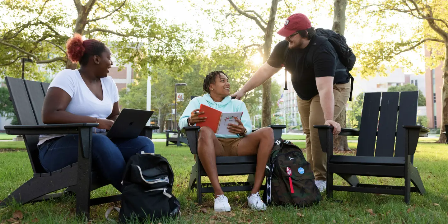 Two student sit studying in adirondack lawn chairs in a shaded part of campus. A third student leads down to talk with one of the seated students.