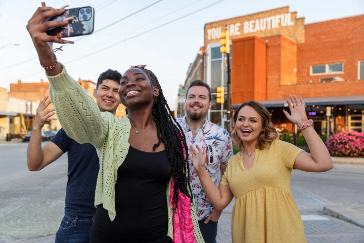 A woman with three other people behind her holds up her phone for a selfie at an Indianapolis location in Fountain Square with a sign on a brick building behind them that reads You Are Beautiful.