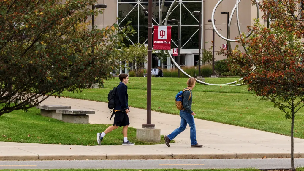 Two students wearing backpacks walk along a sidewalk outside University Library. Red IU Indianapolis banners are seen on light poles and a silver, looping metal sculpture stands nearby.