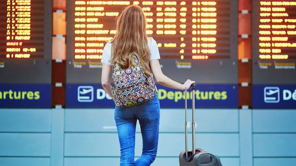 A woman wearing a backpack and hand resting on a roller suitcase next to them stands looking a digital display of airplane departures times.