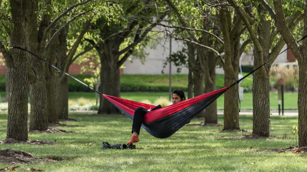 A student sits studying in hammock hung between two trees over grass on the IU Indianapolis campus, one leg dangling out of the hammock.