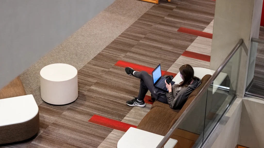 A student sits on an upholstered banquette with side tables in the University Library with laptop on their lap. The student is seen from a floor above, looking down through an open atrium space.
