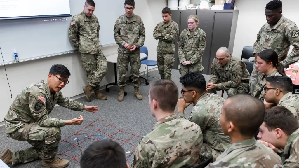 ROTC students in military fatigues gather around an instructor who is pointing at a diagram of colored ropes on the floor.