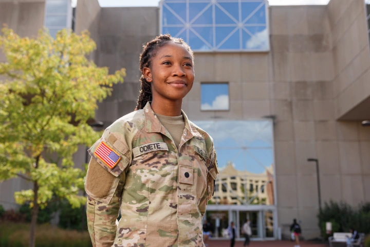 A young woman in an Army uniform stands with her arms clasped behind her in front of University Library, looking off to the side.