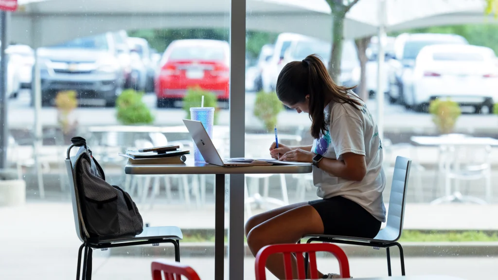 A student sits at a table in the Campus Center food court, with laptop and books on the table and a backpack in the opposite chair. A tree, outdoor seating, and parked cars are seen in the distance through windows.