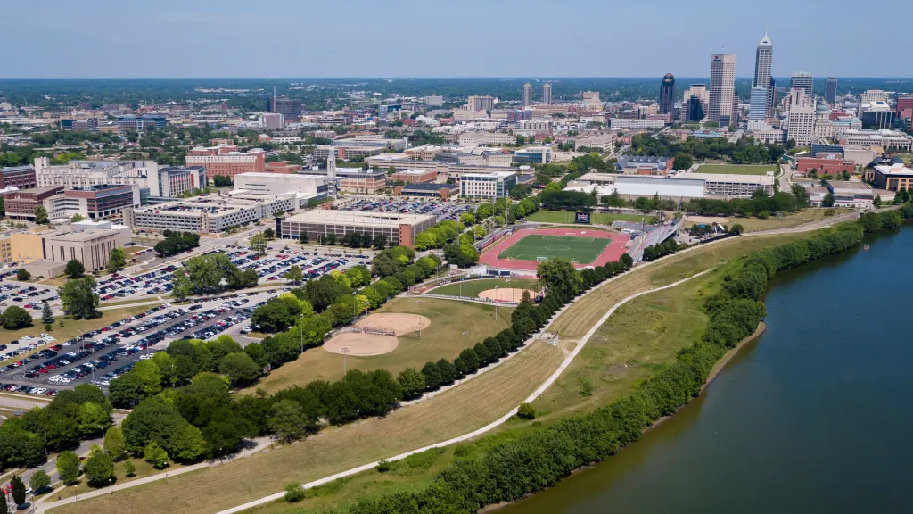 Aerial view of the IU Indianapolis campus including the Campus Center, surrounding buildings and parking lots, Carroll Stadium, the White River flanked by walking paths, and the skyline of downtown Indianapolis in the near distance.
