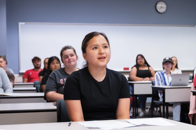 A young brown-haired woman listens intently in a classroom, surrounded by peers.