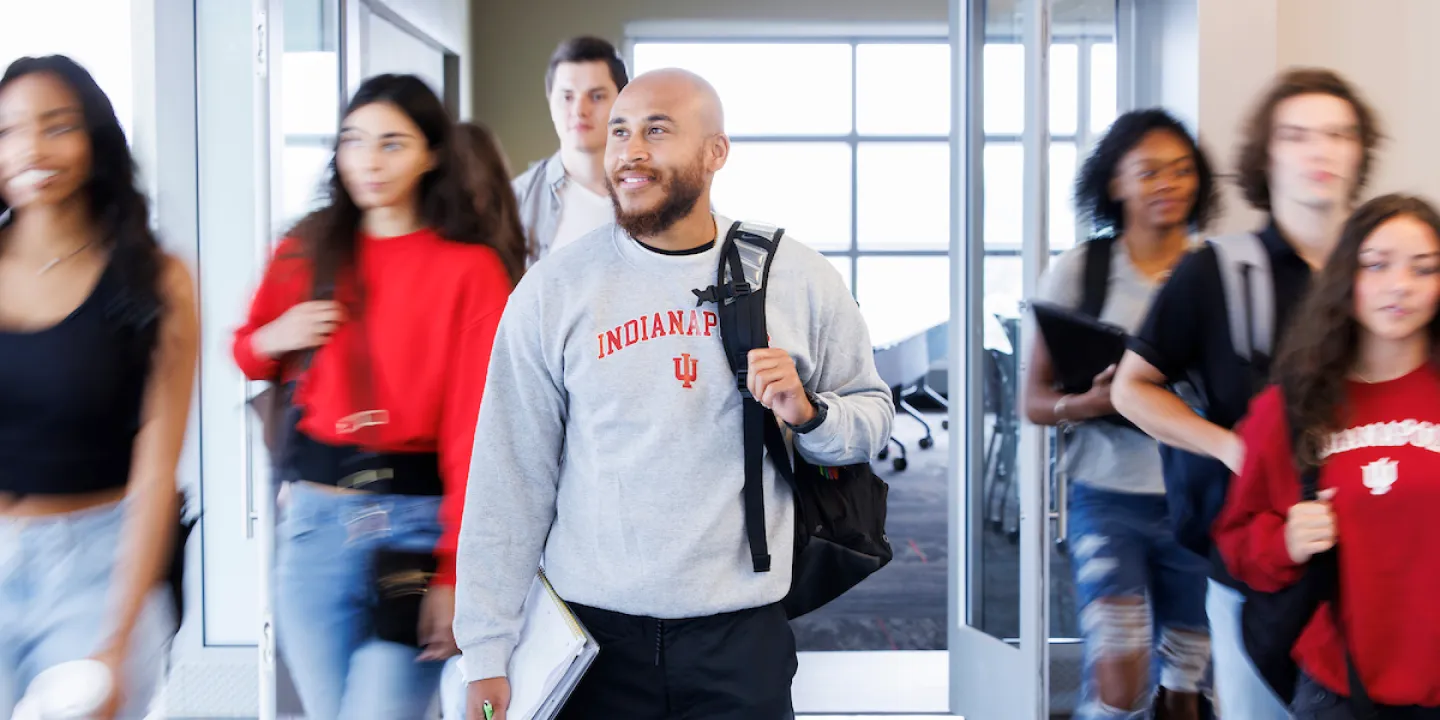 Eight students walk out of a classroom, its glass door open in the background, holding backpacks, books, and laptops. The students are blurred in motion but one student wearing an IU Indianapolis shirt is in focus in the center.