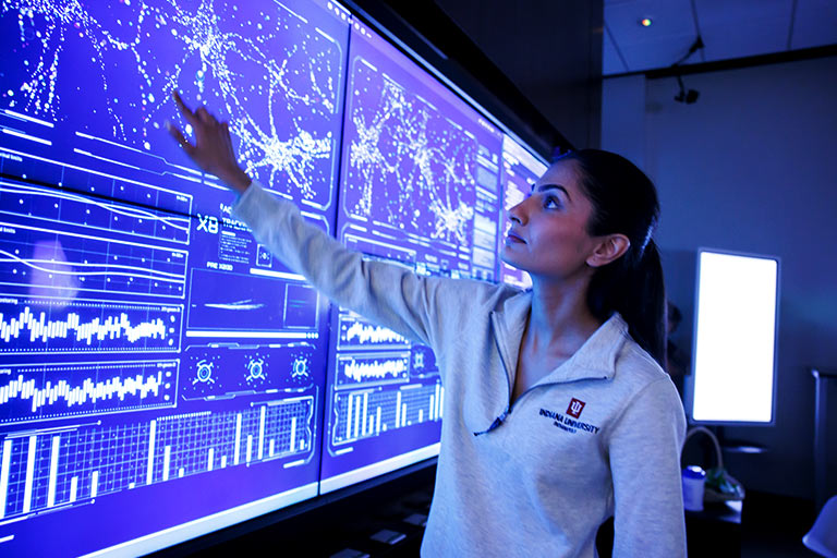 A female student stands in front of a large computer wall, she reaches out her hand to touch the screen.