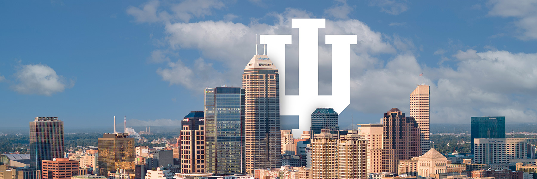 The Indianapolis skyline is pictured on a blue sky day, the IU Trident stands within the city, rising above the buildings.