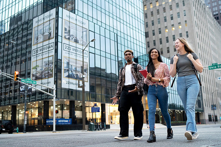 Three students smile and talk while walking through city streets with large glass buildings behind them.