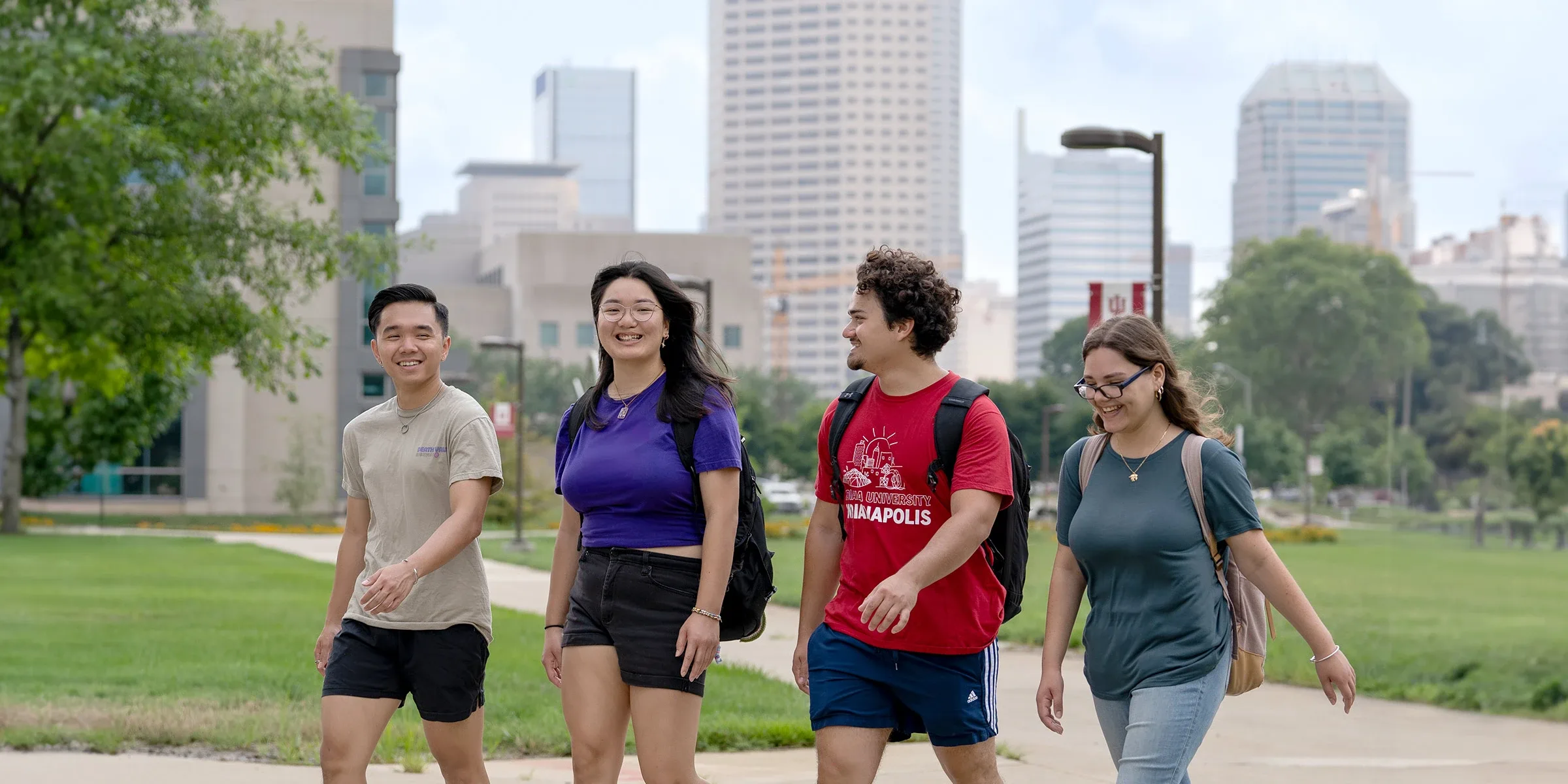 A group of four students walks through campus, tall city buildings are visible in the background.