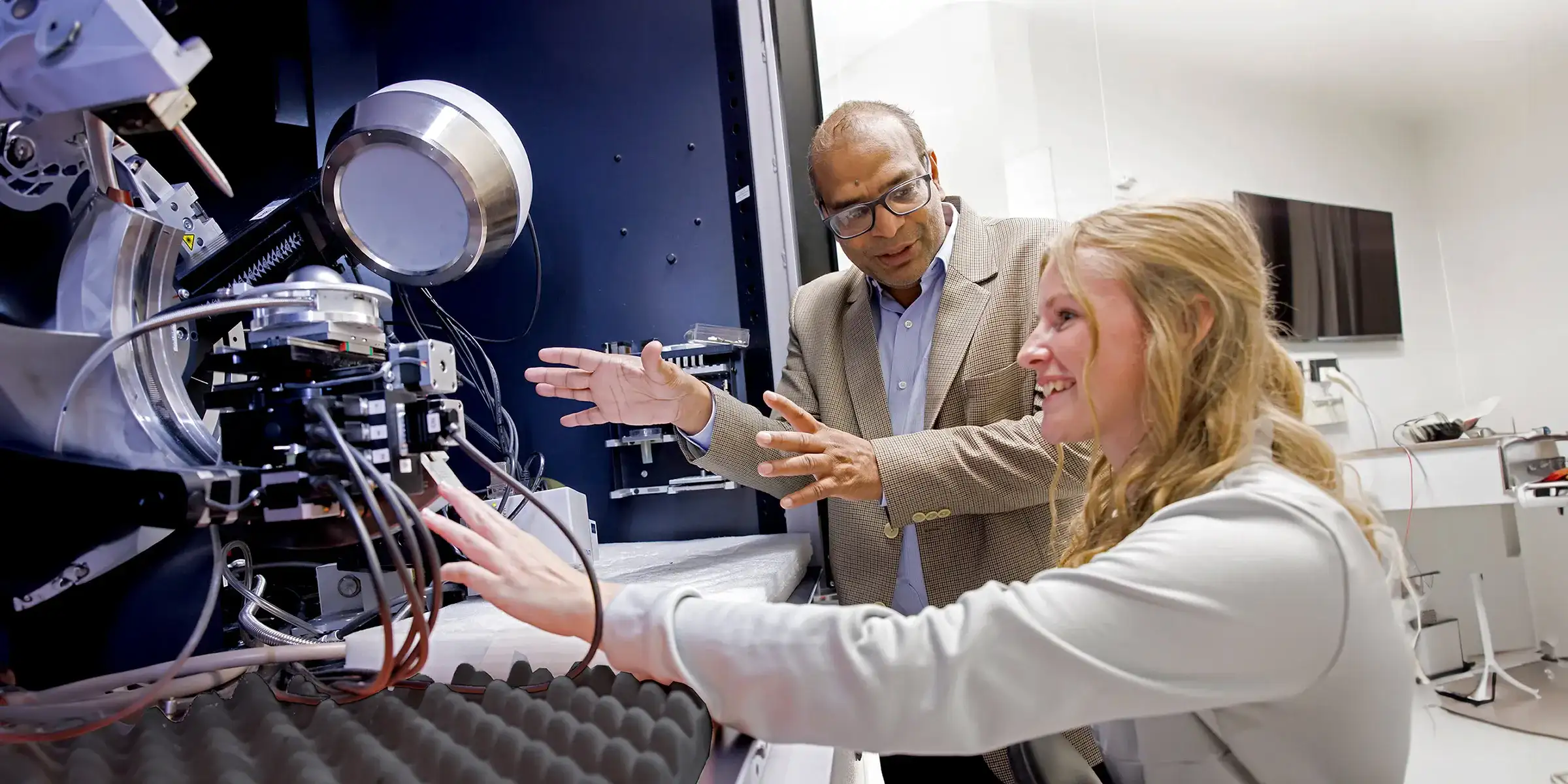 A female student reaches out to touch a piece of lab equipment, her professor stands next to her explaining something.