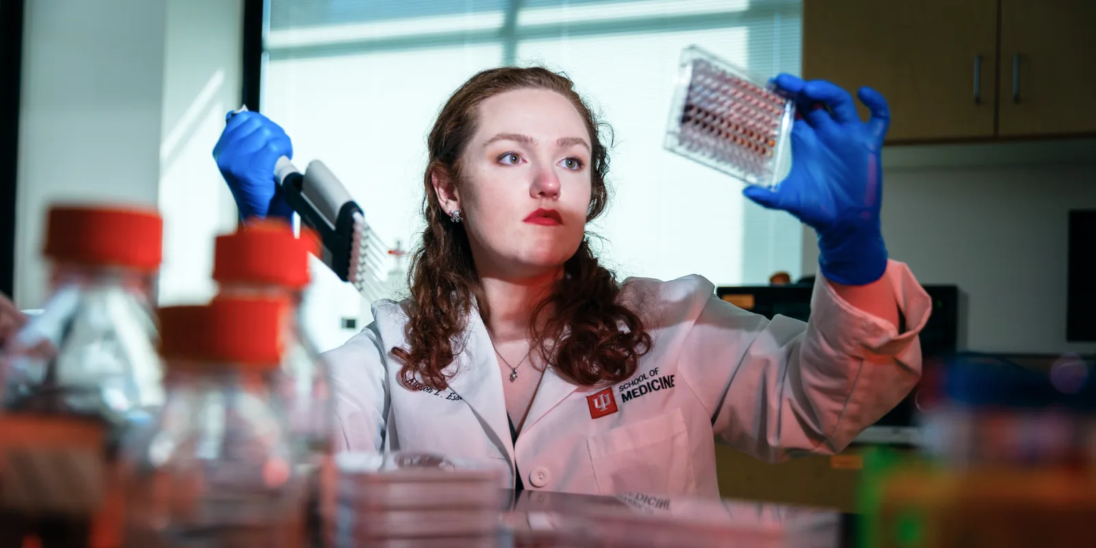 A female student in a lab coat with “IU School of Medicine” logo, wearing blue gloves, examines a lab sample while surrounded by lab equipment and bottles.