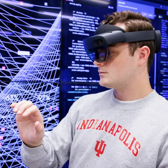 A male student wearing a VR headset and an “Indianapolis IU” sweatshirt interacts with a virtual display, showing intricate patterns and data.