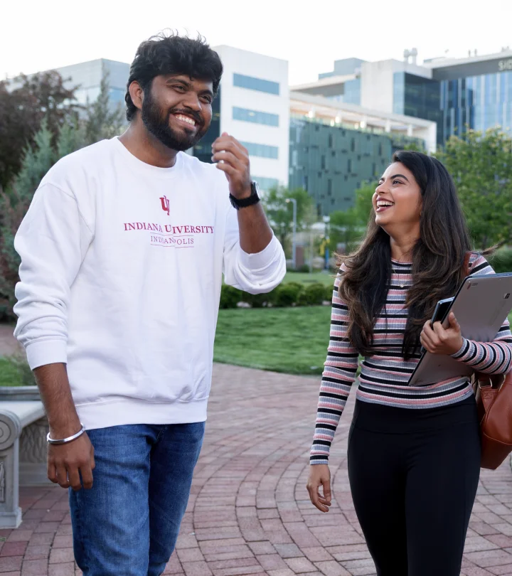 Two students walking outside on a brick pathway, smiling and engaged in conversation. The male student on the left is wearing a white “Indiana University Indianapolis” sweatshirt and jeans, while the female student on the right is holding a tablet and wearing a striped long-sleeve shirt and black pants. Modern buildings and a grassy area are visible in the background.