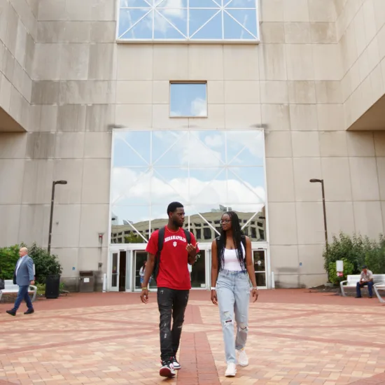 Two students, one wearing a red IU shirt, walk together in a courtyard outside a modern building with large glass windows.