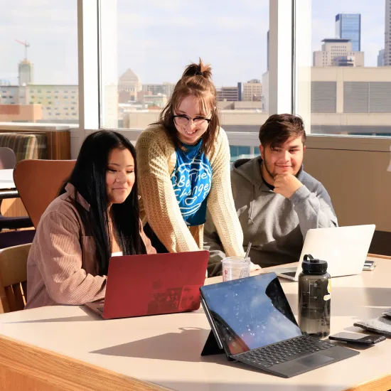 Three students working together on laptops at a table in a bright room with large windows showing a cityscape in the background.