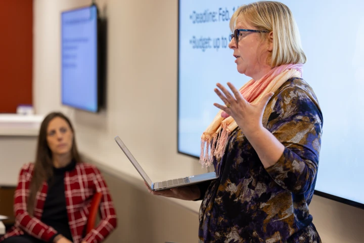 A woman holding a laptop in her hand stands in front of a monitor with words displayed, she gestures with her free hand as she speaks to seated listeners.