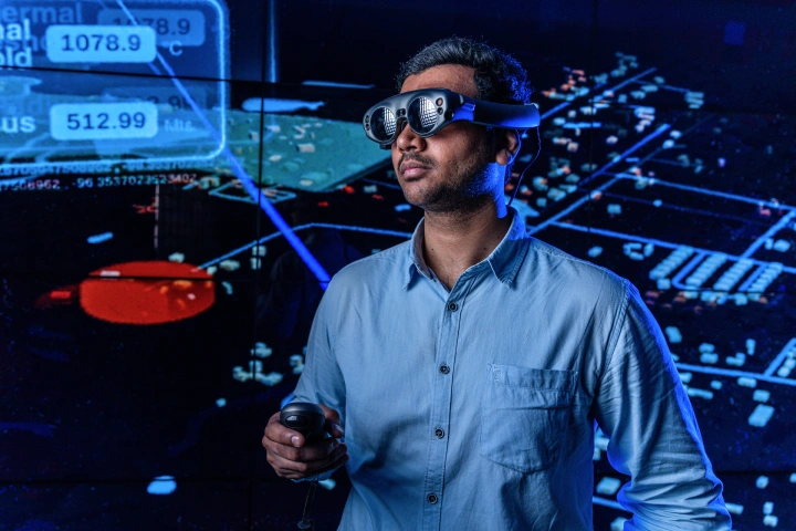 A male student stands in front of a wall screen showing blue lines, figures, and numbers while he wears VR glasses on his face and holds a VR console in his hand.