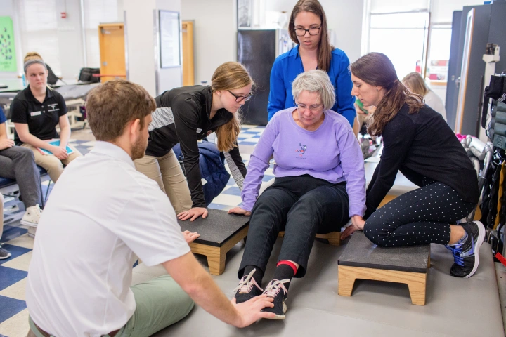 Several students assist woman sitting on the ground with her hands on small step platforms to to a physical therapy exercise.