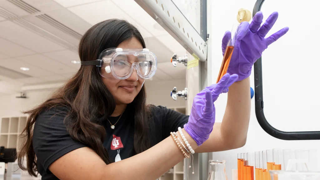 A student wearing safety goggles holds up a test tube of orange solution within a ventilated counter of a lab. Behold her hands are a flask and additional tubes with different mixes of solution.