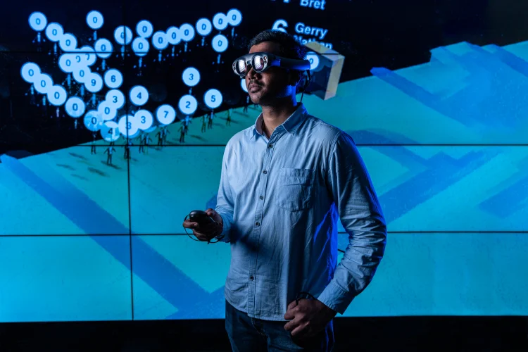 A man stands wearing virtual reality glasses in front of a digital display wall showing several elements including shapes that looks like streets or buildings, and figures of people with numbers hovering above them.