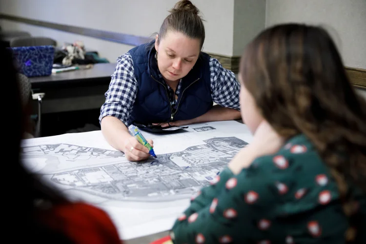 A woman leans over a black and white printed map laying on a table holding a marker in her hand; two other people sit across the table from her.