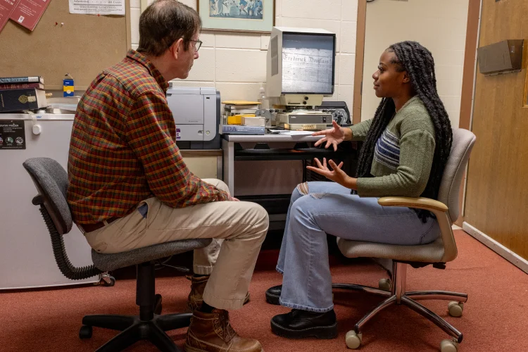 An instructor and a student face each other sitting in rolling in chairs in front of archival equipment for researching historical documents.