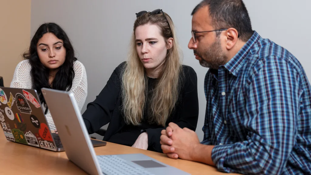 Three people sit at a desk looking at two open laptops in front of them.