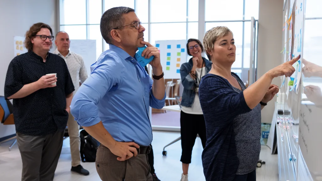 A group of five people gather around a whiteboard in a classroom. One person points at a sticky note on the whiteboard while another thinks, hand on chin.