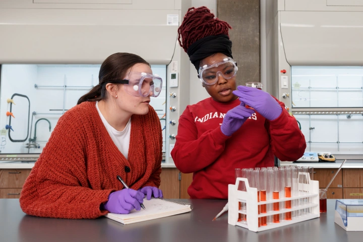 Two female students stand side by side in a chemistry lab, both wearing red shirts, medical gloves, and safety goggles. One holds a test tube of solution and the other takes notes in a notebook.