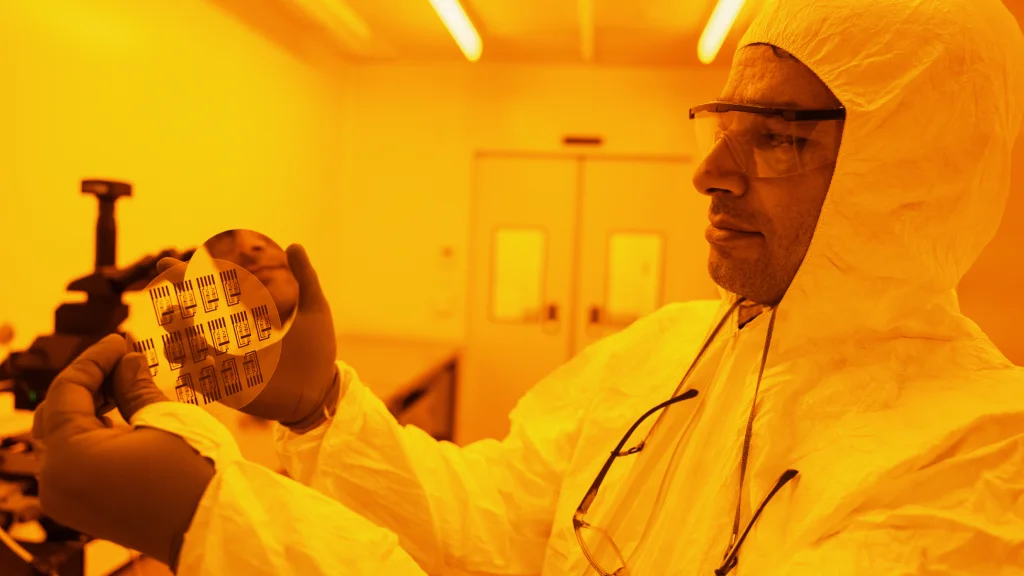 A man wearing safety goggles and protective coverings holds up a circle of chip technology, enlarged by a small mirror or magnifying glass. The man and surrounding lab is caste in a yellow-orange tone of light.