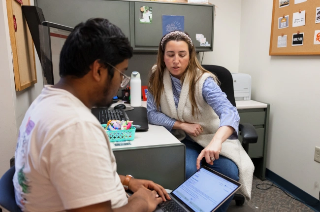 A smart, knowledgeable woman and male student in a small, cozy office look at a laptop together.