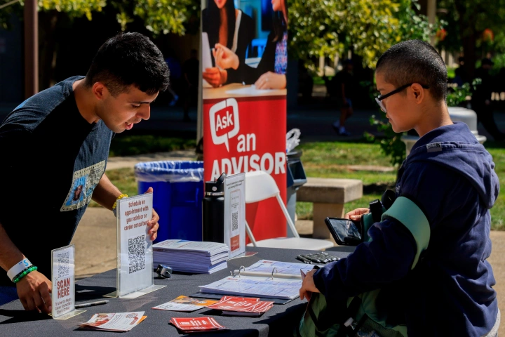 A student stands at an outdoor table with handouts displayed, including a stand-up banner that says Ask an Advisor, as a staff member behind the table points to a scannable QR code displayed.