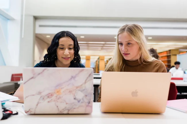 Two students sit side-by-side with laptops open in front of them at a community table in University Library.