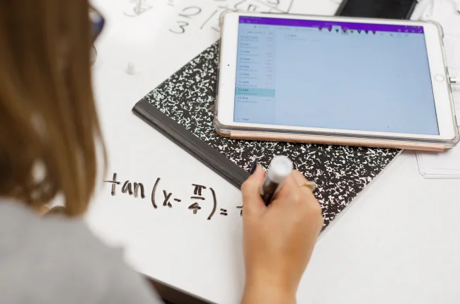 A hand holding a dry erase marker writes out a mathematical formula on a table that also holds a notebook and tablet.