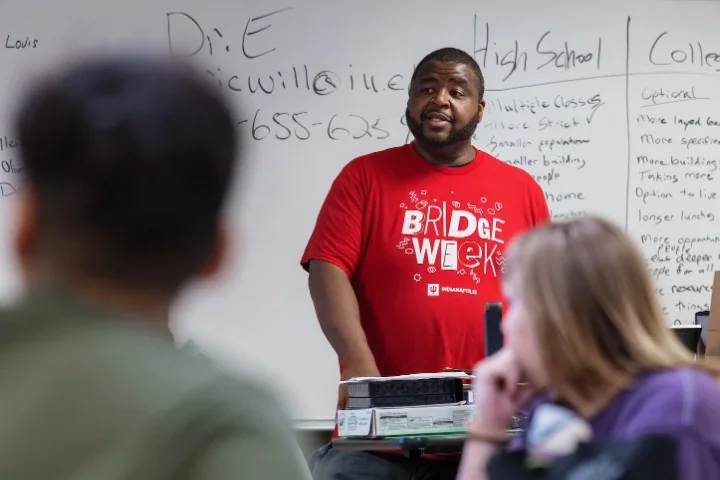 A man in a Bridge Week t-shirt stands in front of a whiteboard talking to a group of students