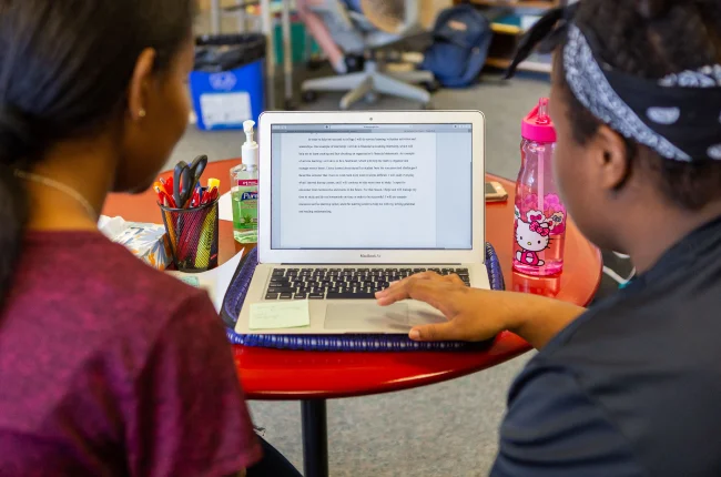 A view over the shoulders of two students looking at a laptop sitting open on a small circular table.